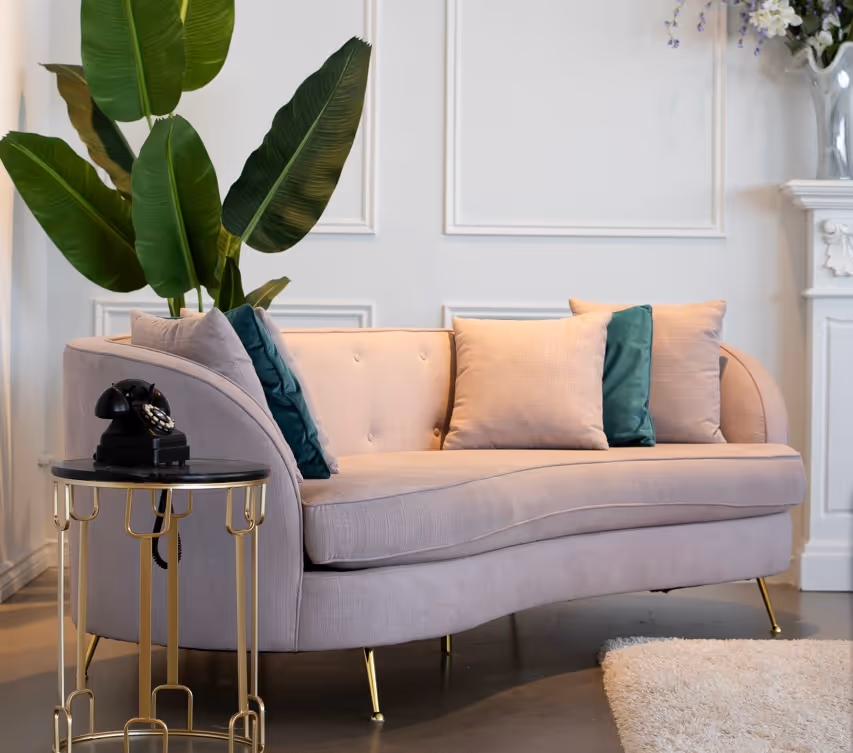 Curved beige sofa with green and beige cushions, gold metal side table with black vintage telephone, green plant in the background in an elegant living room.