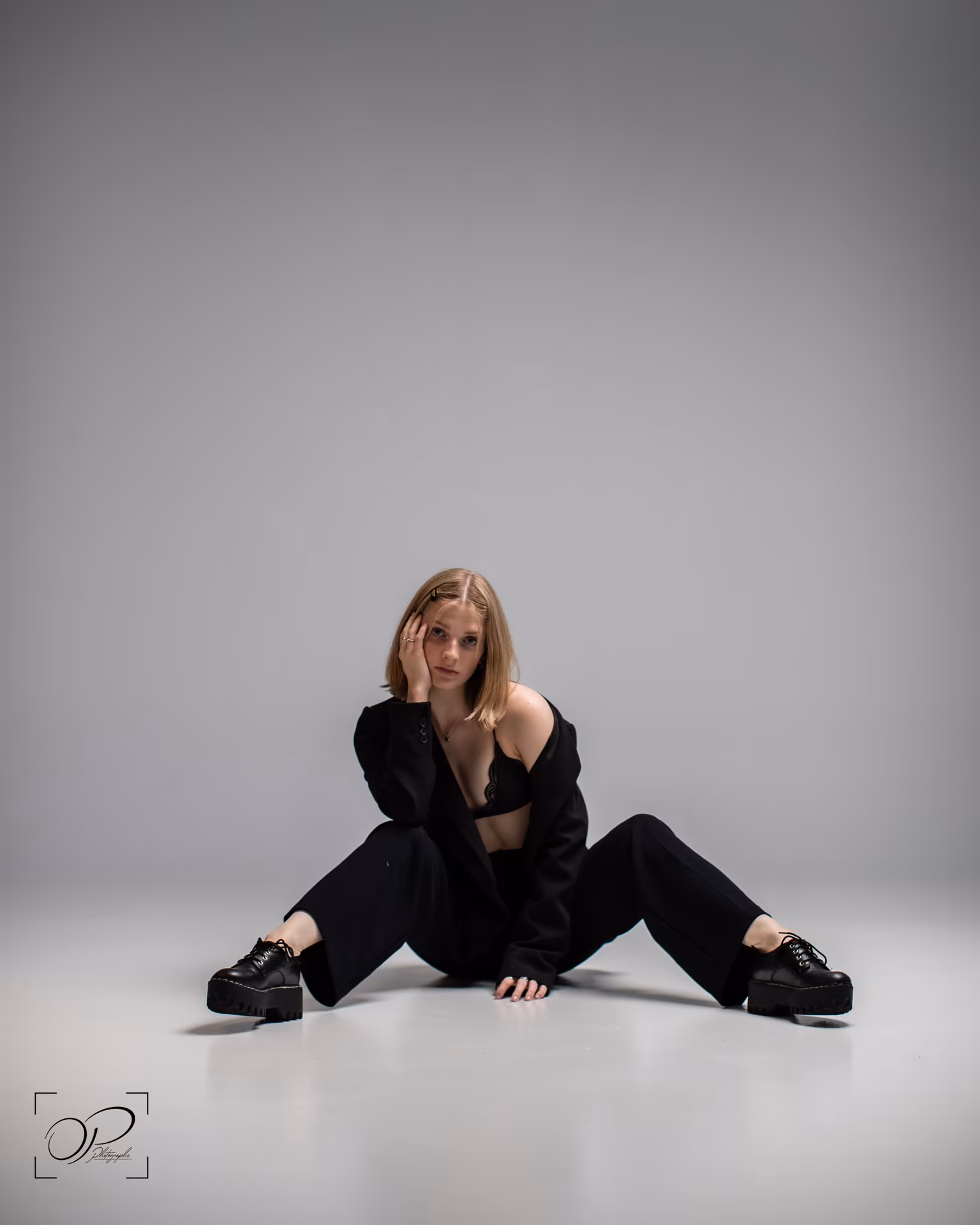 Jeune femme assise au sol, jambes écartées, vêtue d'un blazer noir ouvert sur un soutien-gorge noir et portant des chaussures noires à semelles épaisses.